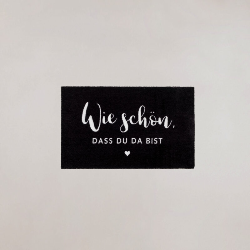 schwarze Fußmatte mit der weißen Aufschrift "Wie schön, dass du da bist"